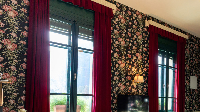 Two windows with dark green trim and red velvet curtains on one wall decorated in black and floral wallpaper.