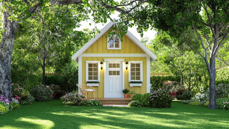 A yellow tiny shed home in the middle of trees on a big lawn.
