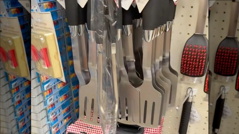 A grill spatula hanging beside metal tongs and grill brushes on the shelf at Dollar Tree.