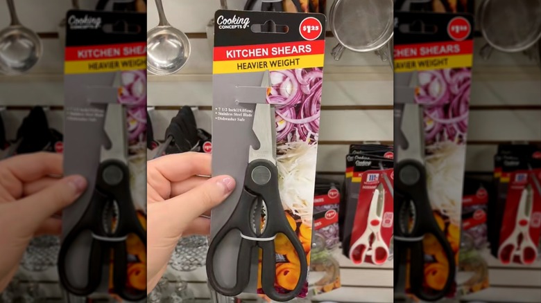 A hand holds a pair of Cooking Concepts Kitchen Shears in front of a display wall at Dollar Tree.