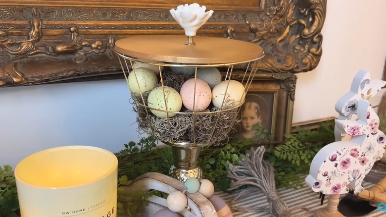 Elevated wire basket on a candleholder with a wood lid filled with decorative eggs and moss