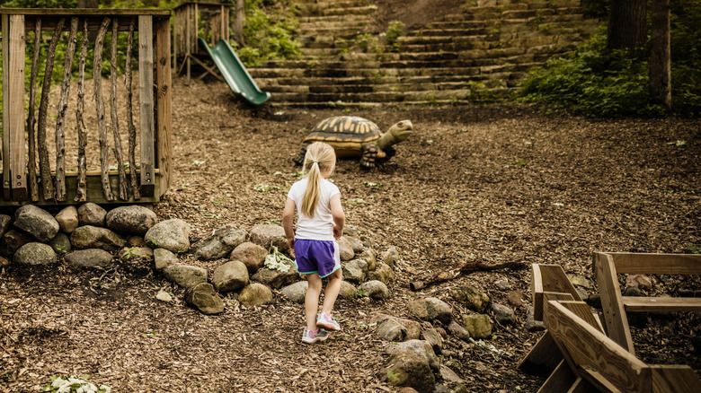 A young girl is seen walking, with her back to the camera, in a wood chip-mulched yard