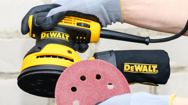 Gloved hands holding highly rated DeWalt sander and sandpaper against white masonry