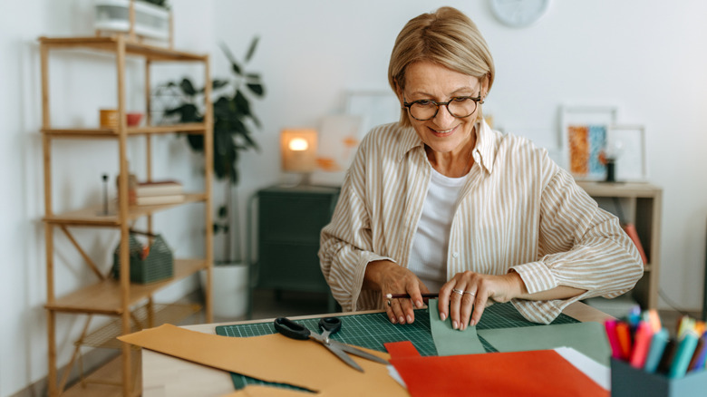 A woman in a striped shirt and glasses cuts out colorful paper in her home.