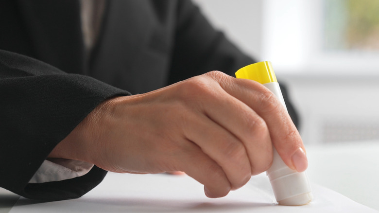 A person wearing a black jacket presses a glue stick onto a piece of paper.
