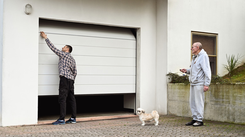An old man with a white dog in front of him pressing a garage door remote in front of the garage while a younger man inspects the door