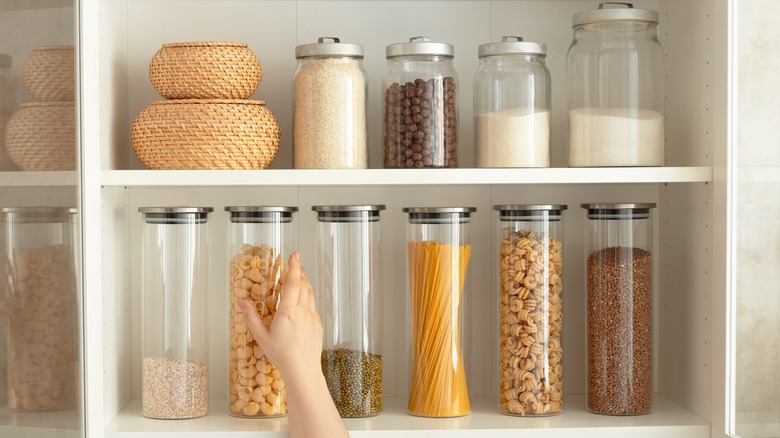 Woman reaching for a glass storage container filled with pasta from a stocked pantry