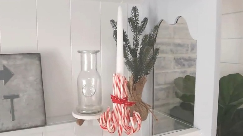 White taper candle with candy canes forming a holder, sitting on a table next to a pine tree and glass jar