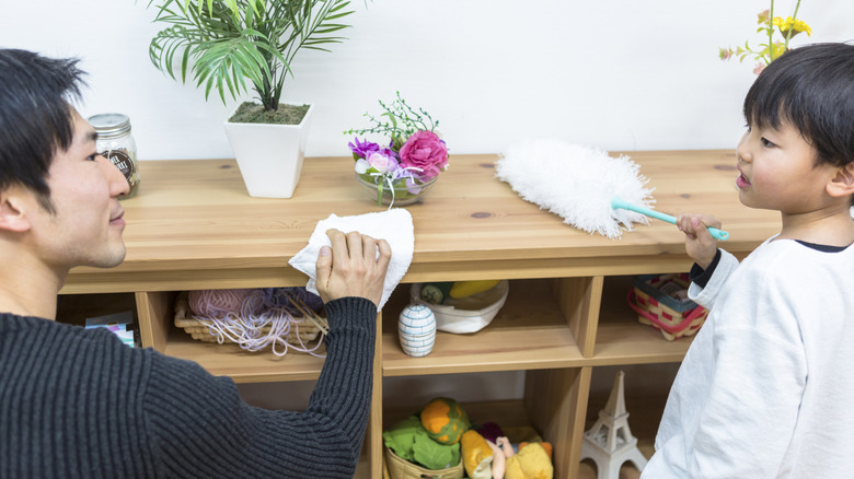 Parent and child wiping down a shelf with toys and craft supplies on it
