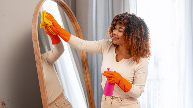 Woman cleaning a full-length freestanding mirror in a bedroom next to a window