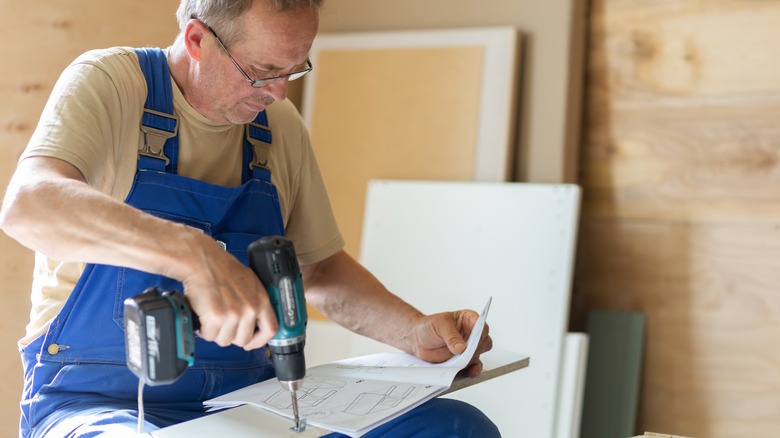 An older man in a beige shirt and blue overalls using a drill