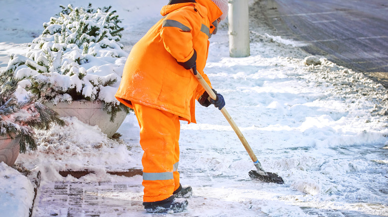 Person wearing an orange coat and snow pants using an ice scraper tool to remove ice from sidewalks