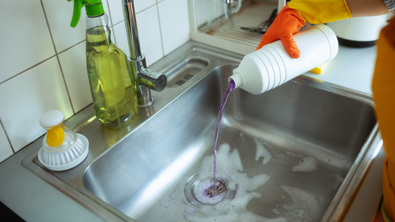 A gloved hand pouring drain cleaner into a stainless steel kitchen sink