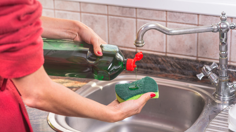 A person squeezing green dish soap from a bottle onto a green and yellow sponge