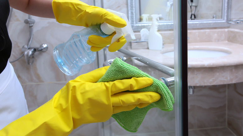 Hands with yellow rubber gloves using a microfiber towel and spray bottle on a glass shower.