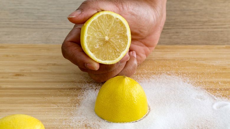A hand holding half a lemon as the other half rests on a wood cutting board in baking soda.