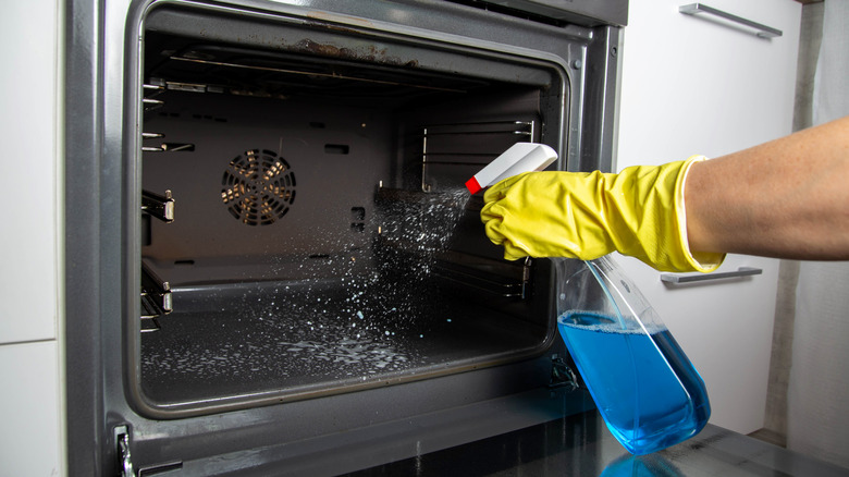 A hand with a yellow rubber glove using a spray bottle on the interior of an oven