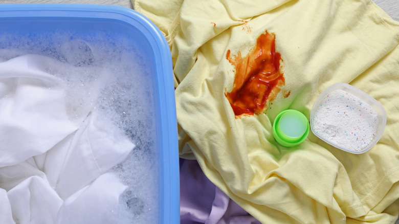 A wash basin with white sheets beside a yellow shirt with a ketchup stain