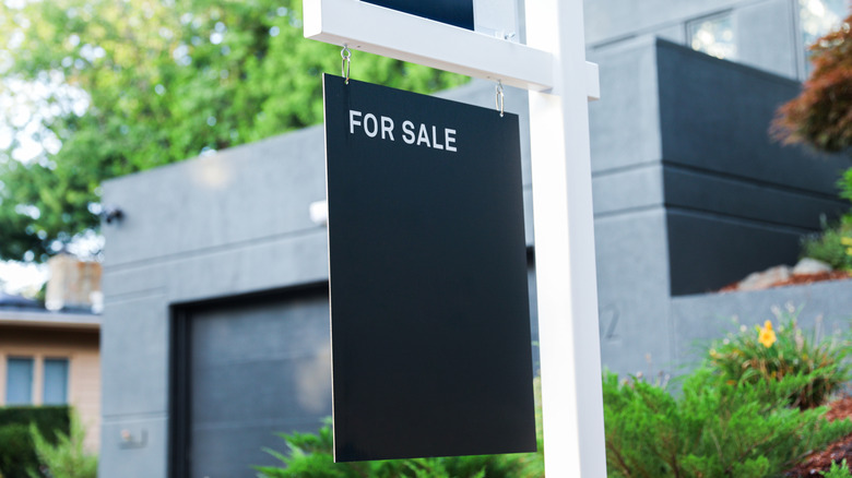 A black for sale sign hanging on a white post outside of a modern-style home with a black garage door