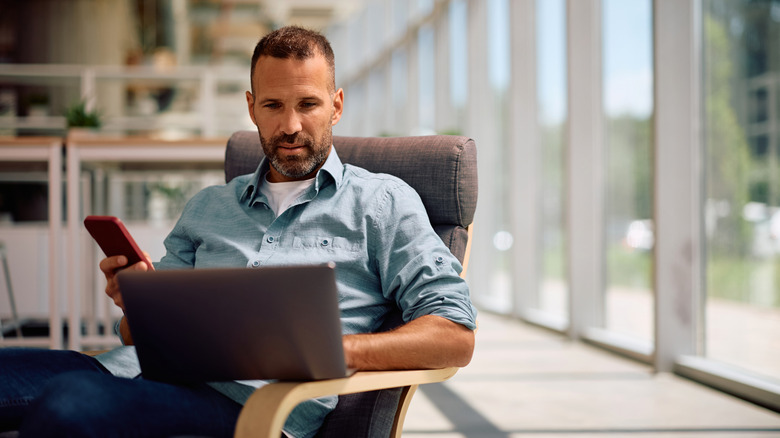 Man in an armchair looking at a computer on his lap and holding a phone
