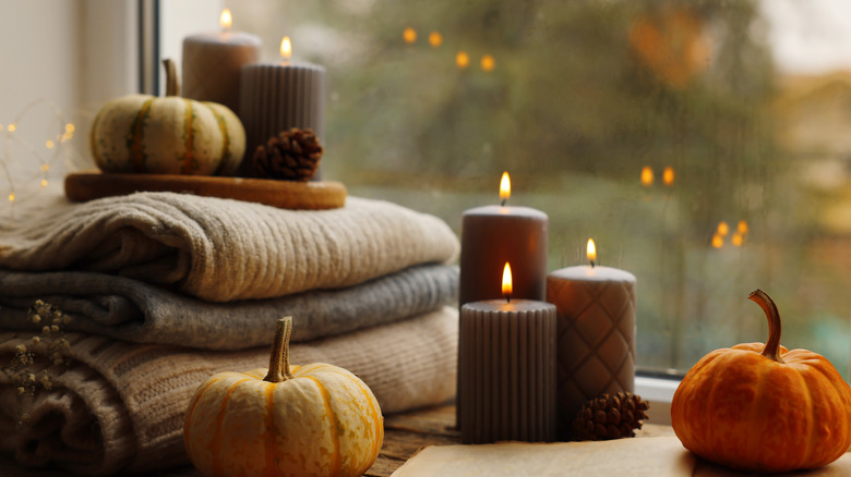 Stacks of blankets, candles, acorns, fairy lights, and pumpkins near a window on a cloudy or overcast day