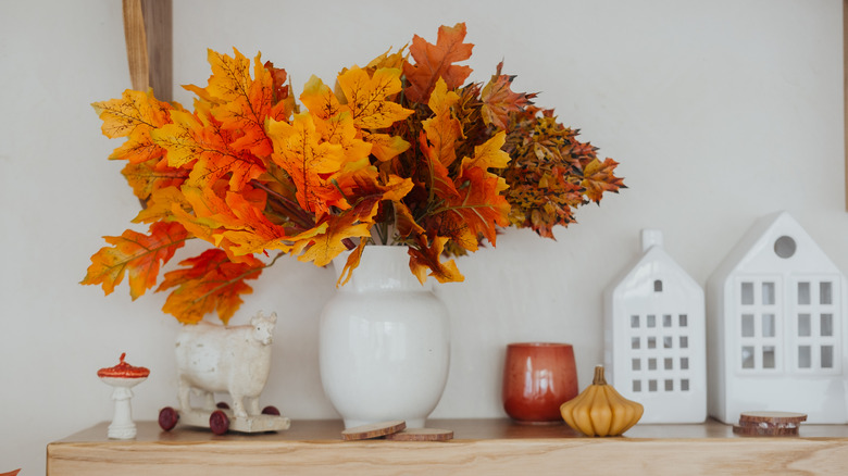 A white vase with fall foliage coming out of it on a shelf with other small fall and white decorations