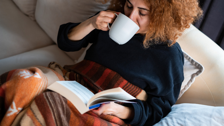 A woman sittin gon the couch with a white mug, a book, and a fall-colored throw blanket