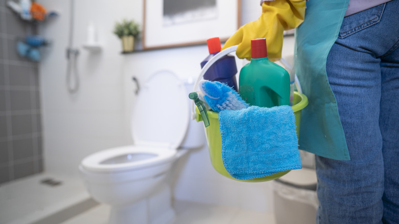 Person standing in a bathroom holding a bucket of cleaning supplies