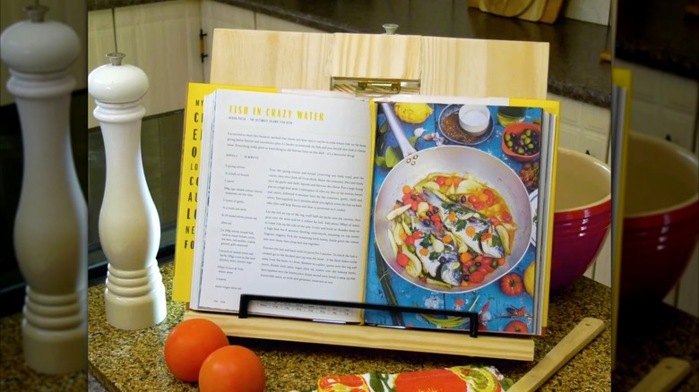 Cookbook resting on a wood stand with a pepper mill, bowl, tomatoes, and spoon on a counter
