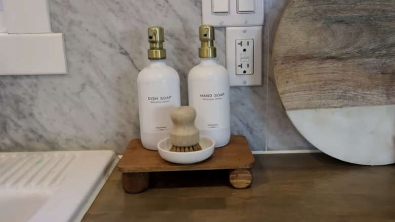 Dish soap, hand soap, and a scrub brush sitting on a small wood shelf near a sink in a kitchen