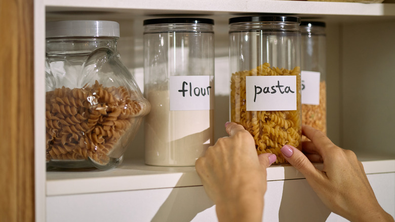 Hands placing a jar labeled "pasta" on a white shelf next to other labeled jars