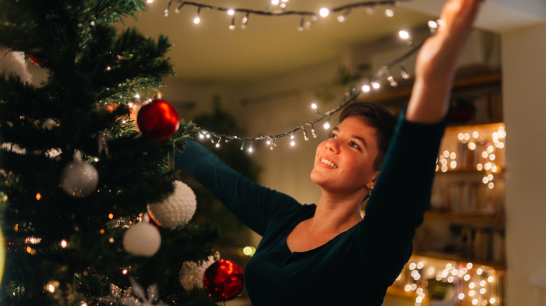 A smiling woman is draping a string of lights on her Christmas tree