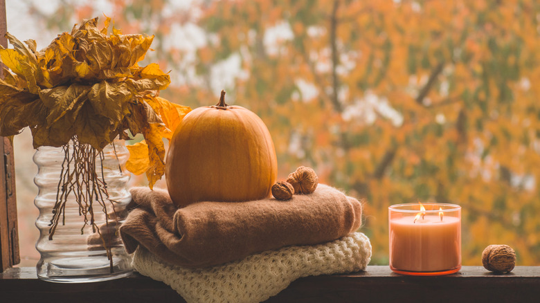 Thanksgiving/Fall decorations stacked on a windowsill, including old leaves, comfy blankets, walnuts, a pumpkin, and a lit orange candle