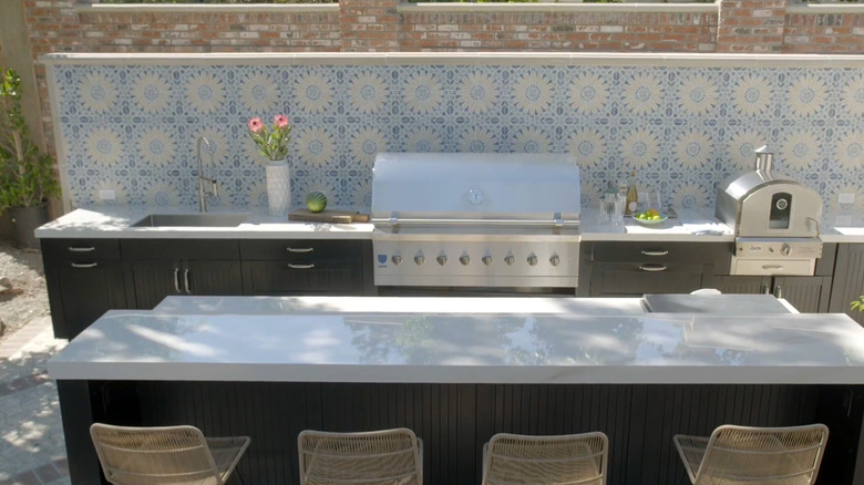 An outdoor kitchen with grill, bar and seating, before a blue and white floral tile backsplash