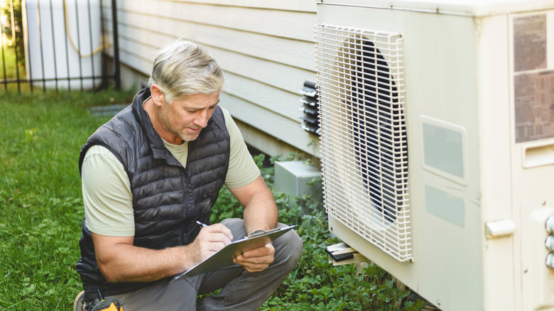 A man working on a clipboard next to a heat pump outside a home