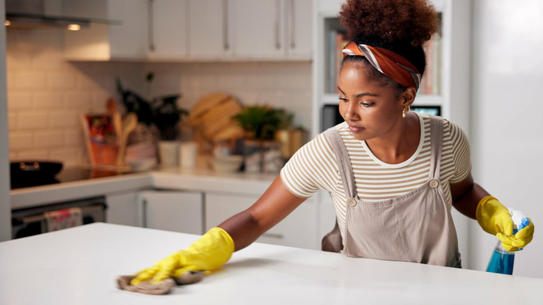 Woman cleaning kitchen island countertop with yellow rubber gloves, a cloth, and spray bottle