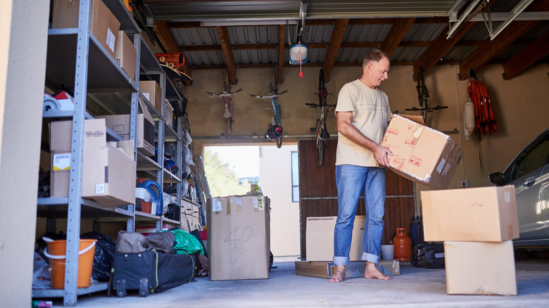 Man holding a box in a garage with additional boxes sitting on the floor and other storage items on metal shelves
