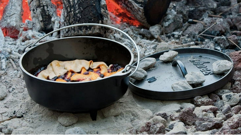 A black Dutch oven sitting amidst ashes of a cooking fire
