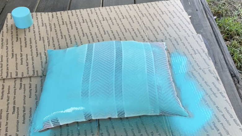 A striped outdoor cushion spray painted turquoise sits on cardboard on a wooden deck.