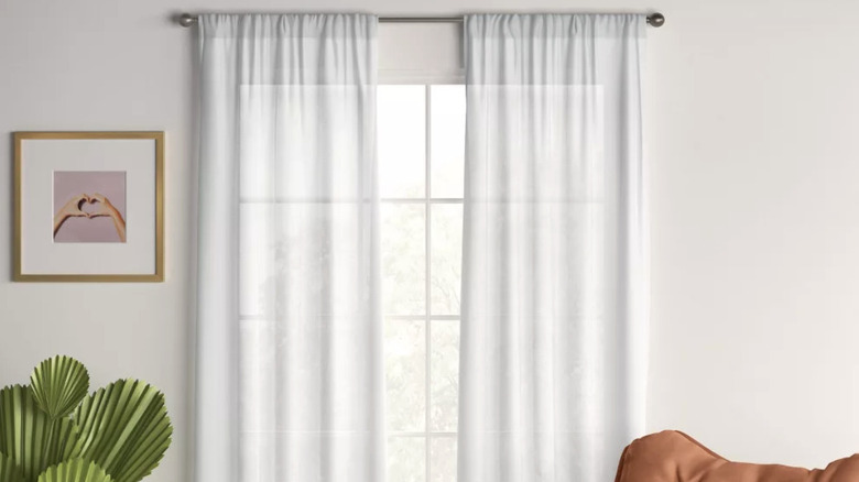 White sheer curtains on a window on a white wall with a plant, art, and chair