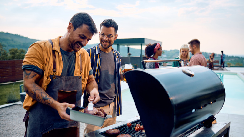A man laughing while tending to the grill, talking to a friend, with others enjoying the party on the patio in the background