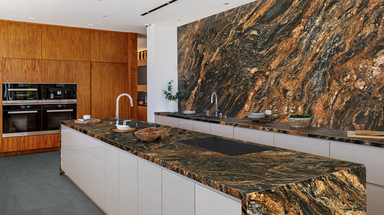 A kitchen with dark backsplash and counters and a natural wood paneling holding double ovens in the background
