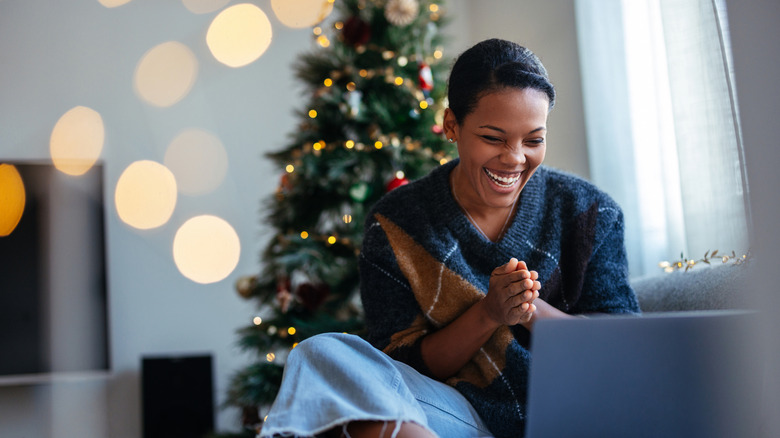 A woman sitting in front of a lit Christmas tree smiles while looking at her laptop