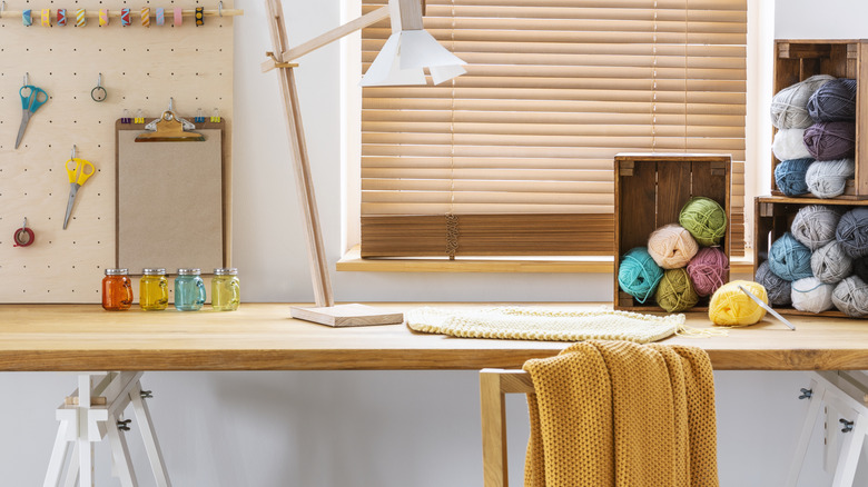 A desk with balls of wool, scissors hanging on a board, lamp on the desk, and chair with yellow throw draped over it.