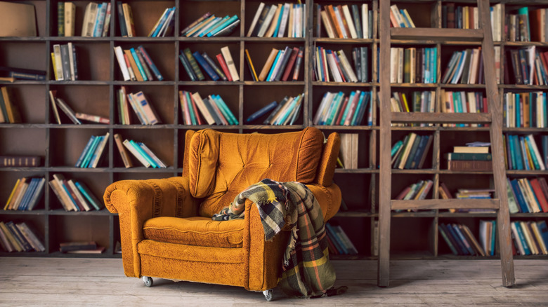 An orange velvet armchair with blanket in front of bookshelves with multiple books on each shelf.