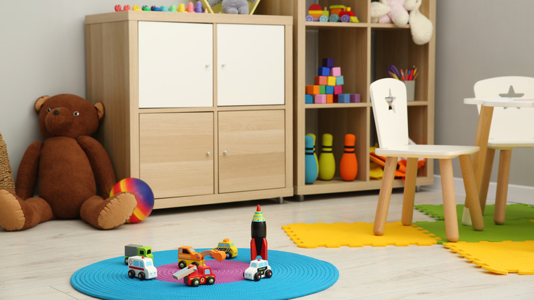 A playroom with open shelving and colorful toys with a big brown bear on the floor and two small chairs on a colorful mat.