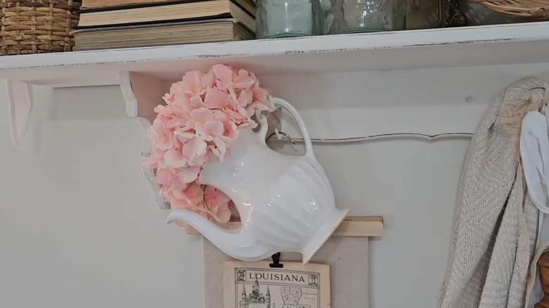 A white pitcher holding pink flowers is hanging from a hook