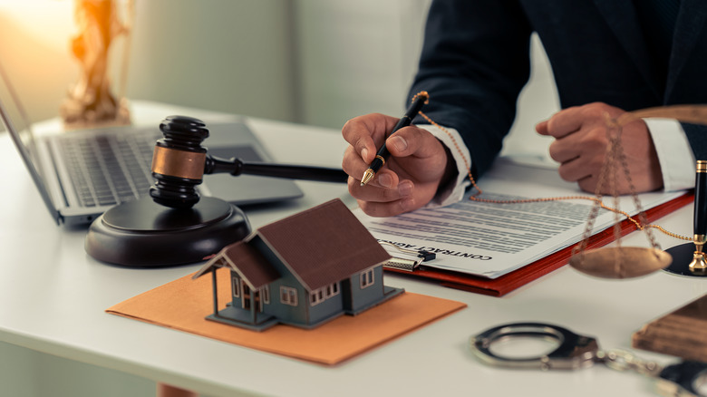 Cluttered desk in a law office includes a laptop, a gavel, a tiny model of a house, handcuffs, the scales of justice and a person holding a pen over paperwork labeled "contract."
