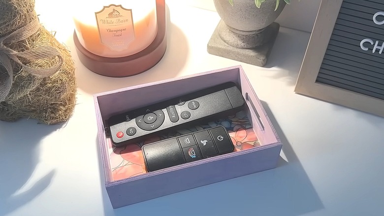 Painted wood tray with a decorative image on the bottom holding remotes next to a candle and decor