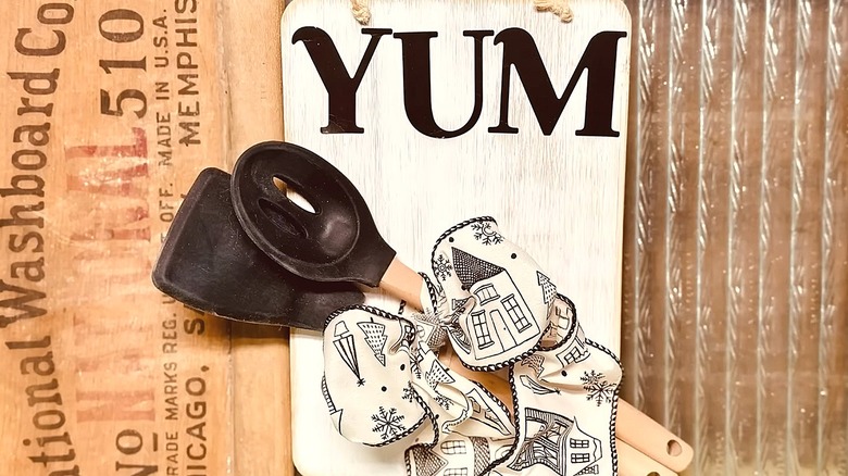 Wooden kitchen sign made with a cutting board, utensils, and a bow attached